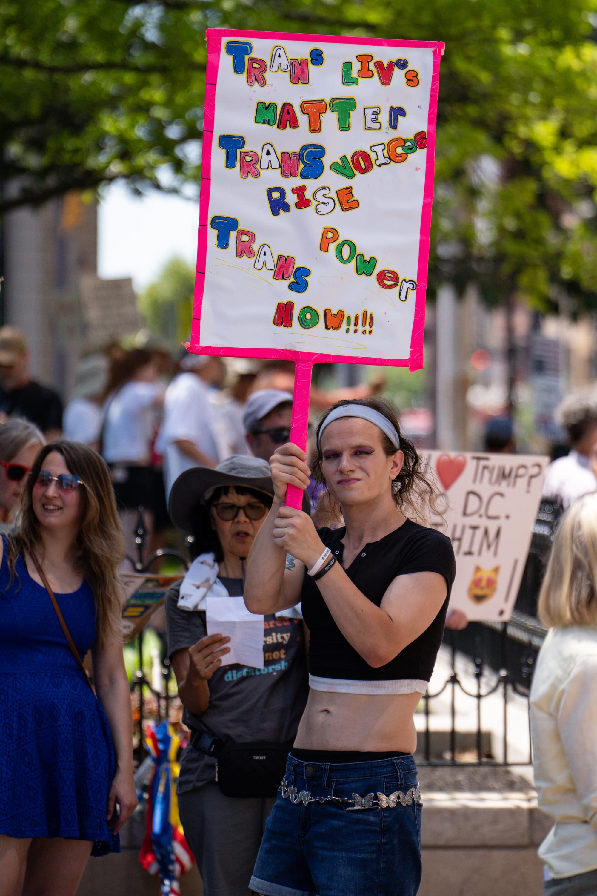 A marcher holds up a hand-painted sign reading 'Trans lives matter, trans voices rise, trans power now!!!'
