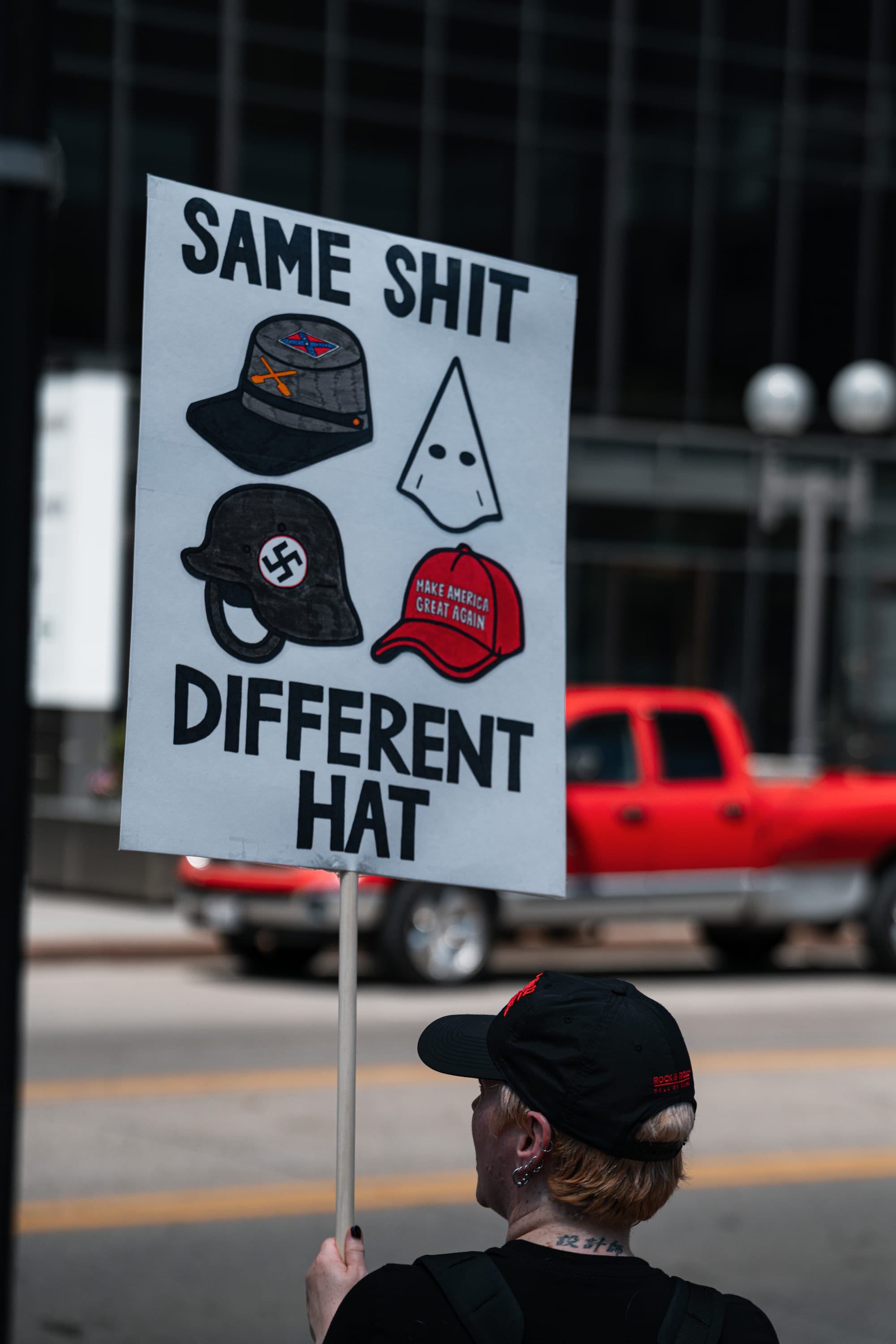 A hand-drawn sign reading 'Same shit, different hat', showing a swastika helmet, a Klan hood and a red MAGA cap, held up beside a downtown street.