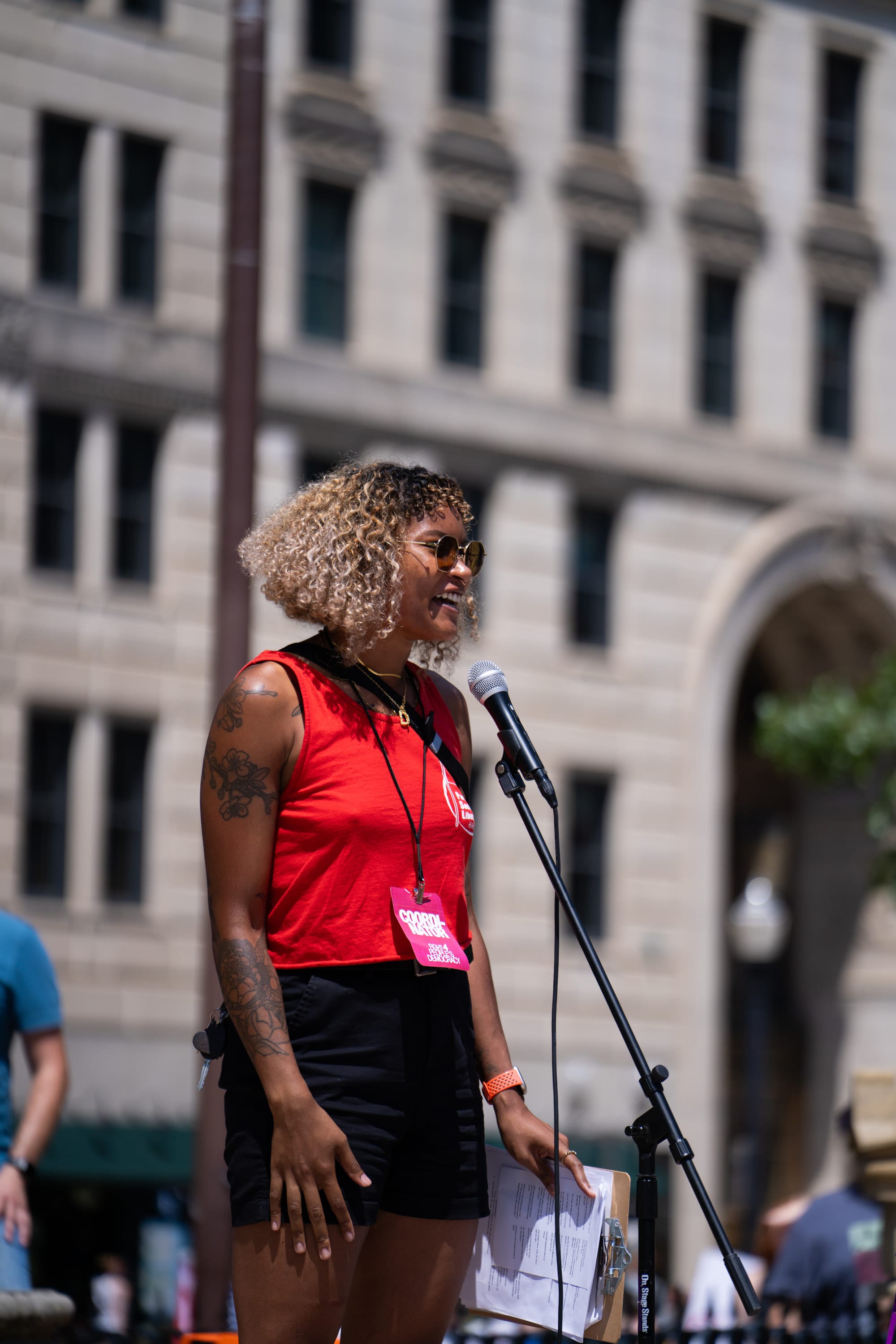 A speaker in a red top smiles at the microphone in front of a stone office building.