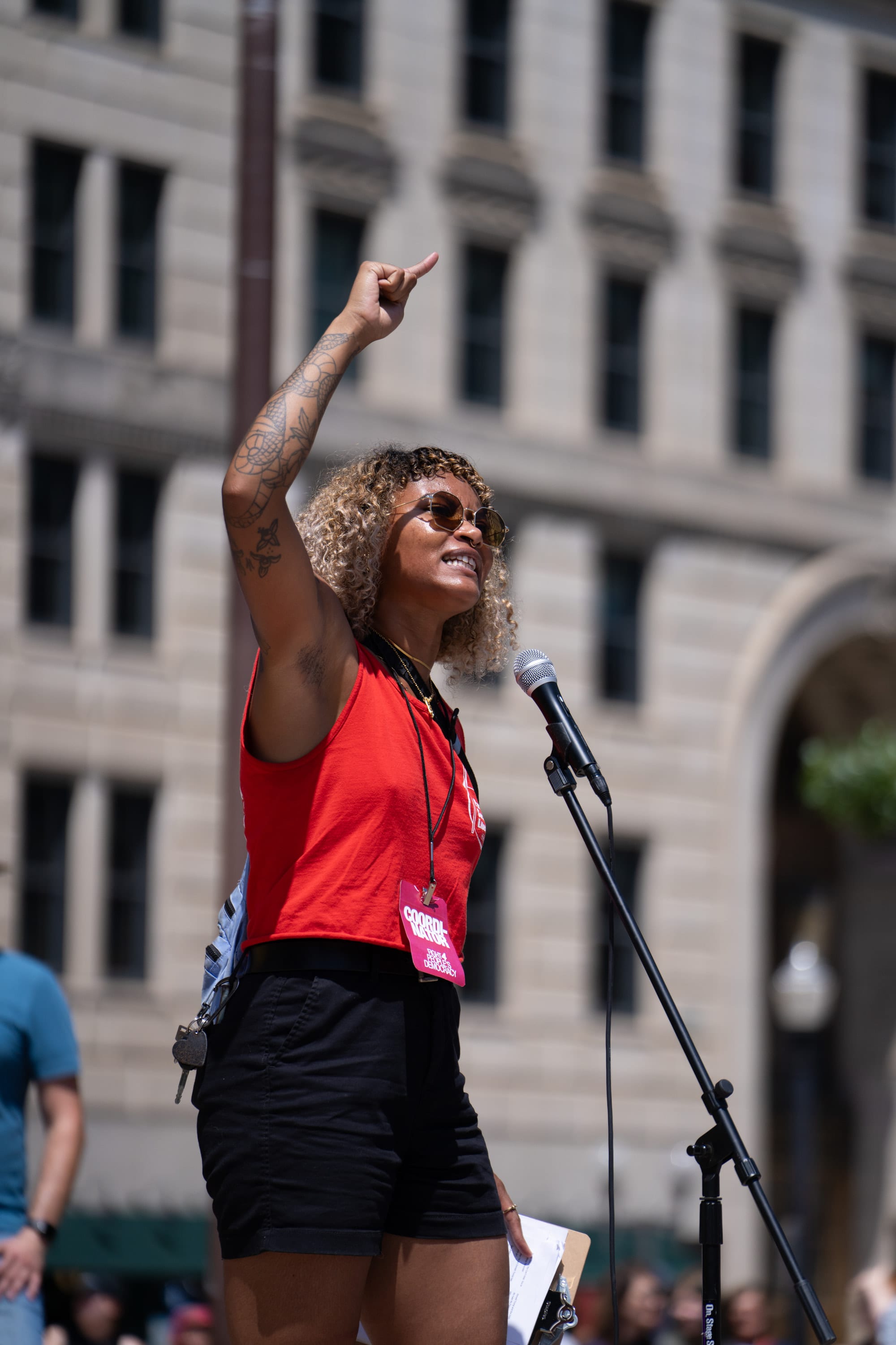The same speaker points skyward mid-speech, the microphone on its stand beside her.