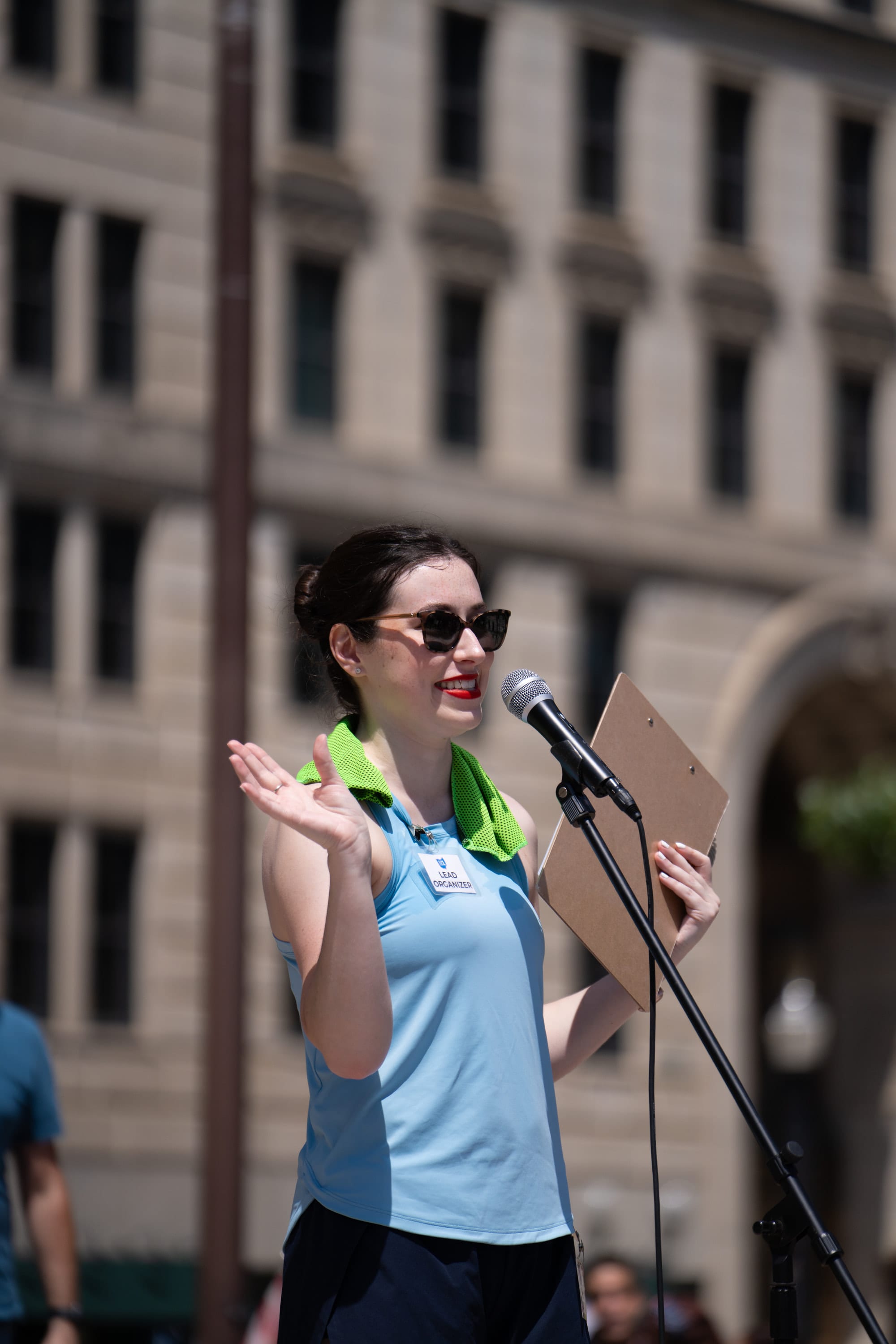 A speaker in a light blue shirt gestures with an open hand at the microphone, notes in her other hand.