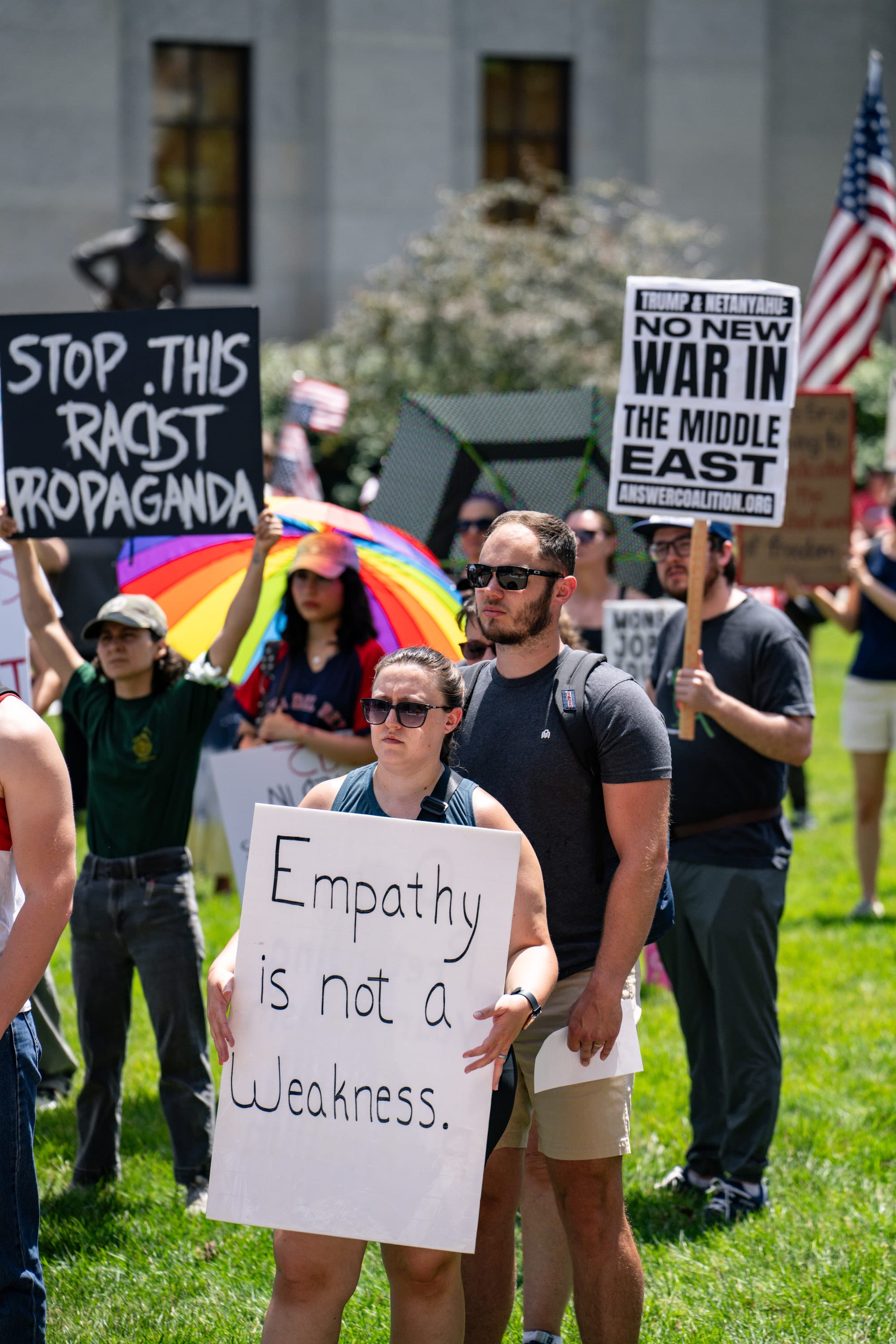 Protesters on the lawn hold signs reading 'Empathy is not a weakness', 'Stop this racist propaganda' and 'No new war in the Middle East', a rainbow umbrella among them.