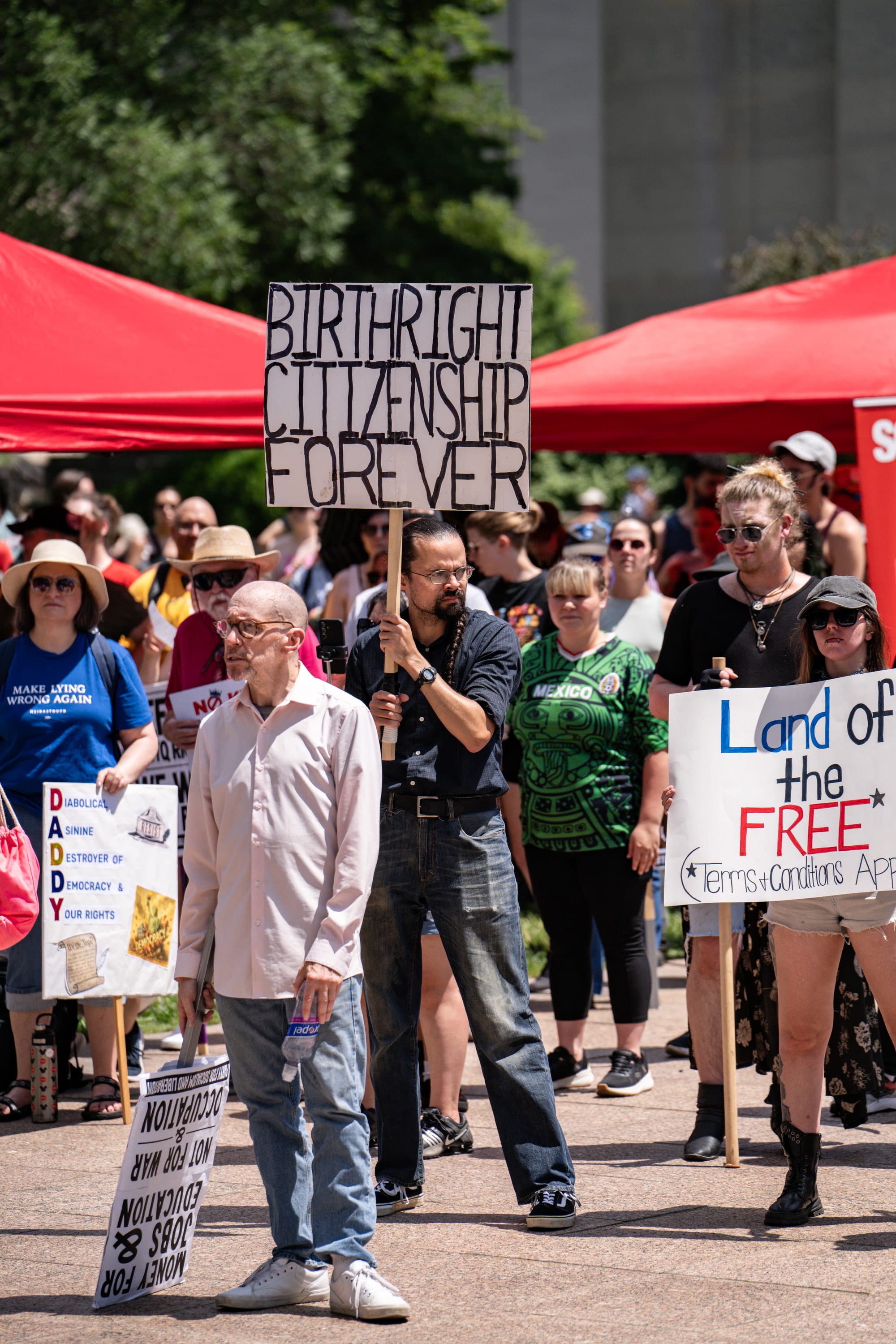 An older man walks past red canopies and a crowd holding 'Birthright citizenship forever' and 'Land of the free' signs.