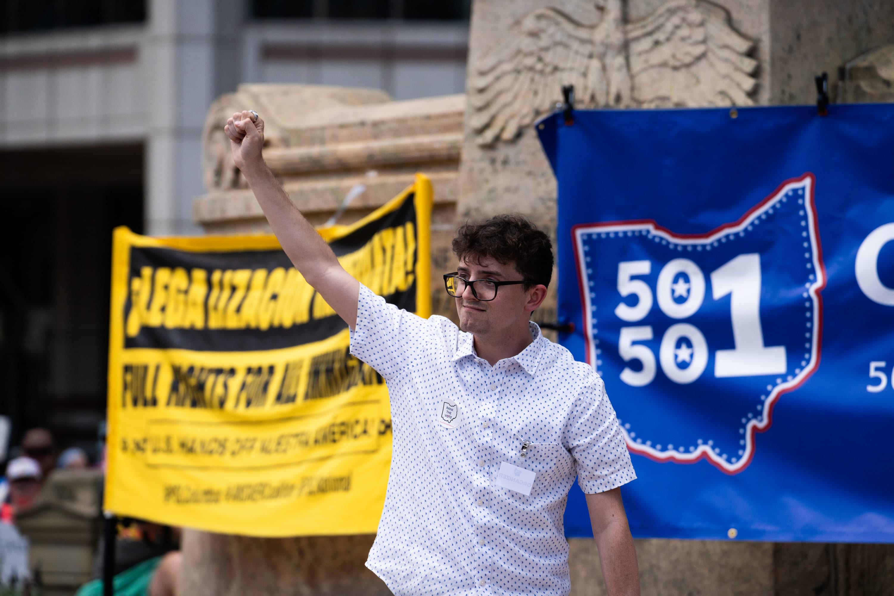 A young man raises a fist beside a blue 50501 Ohio banner and a yellow banner calling for full rights for immigrants.