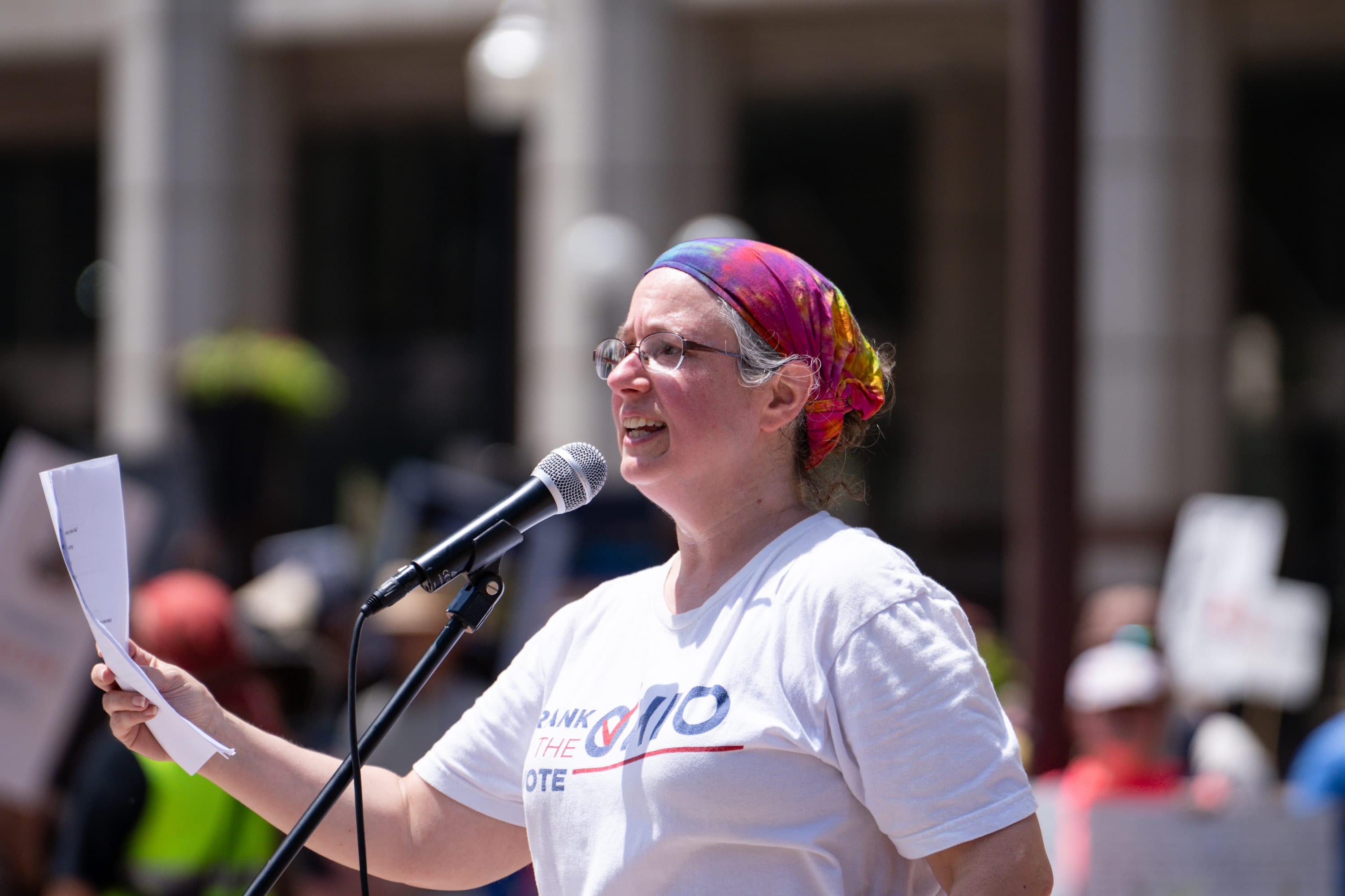 A speaker in a rainbow headscarf reads from a sheet of paper into the microphone.