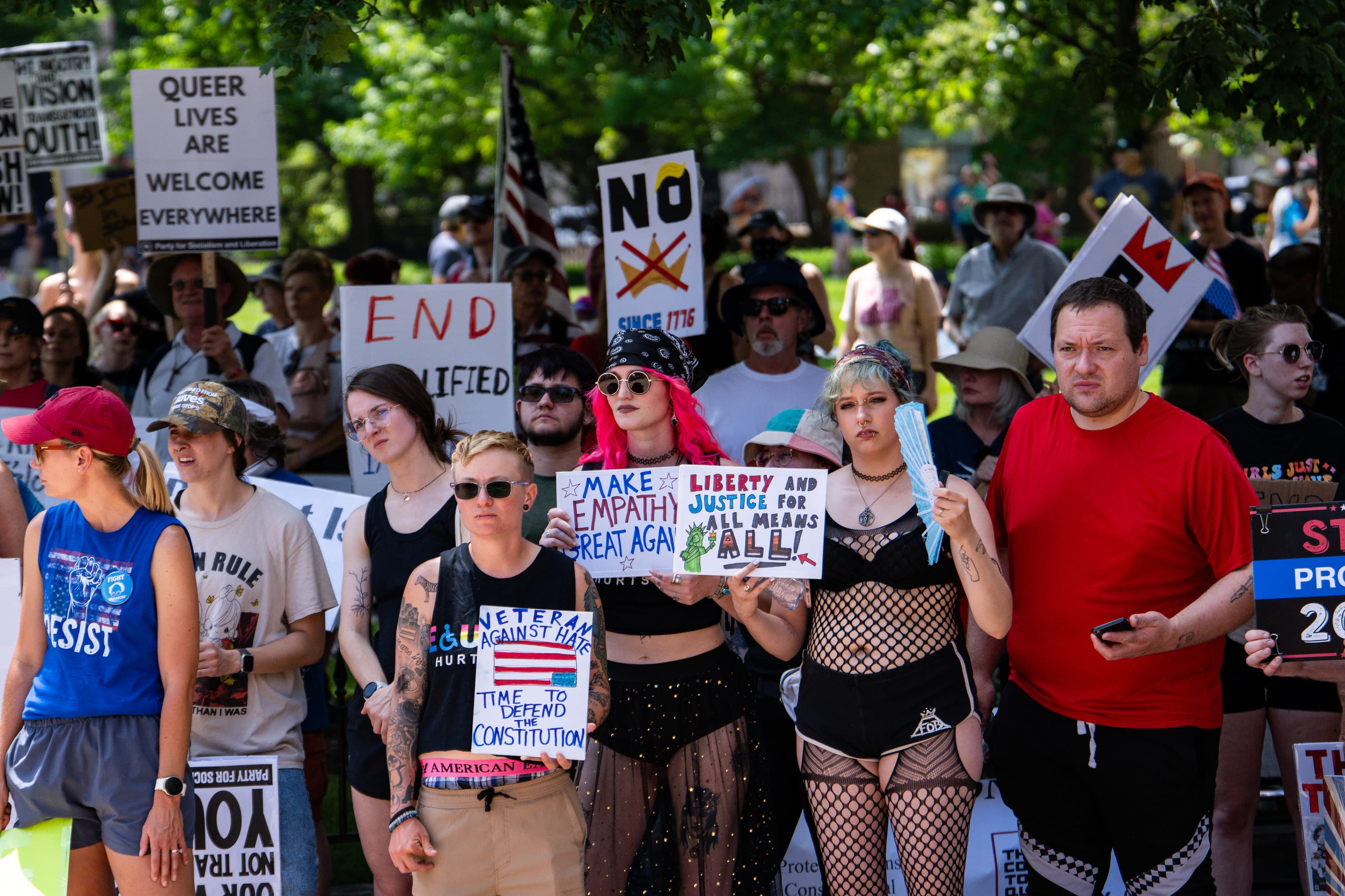 A crowd packed with signs under the trees: 'Queer lives are welcome everywhere', 'Make empathy great again', 'Liberty and justice for all means all'.