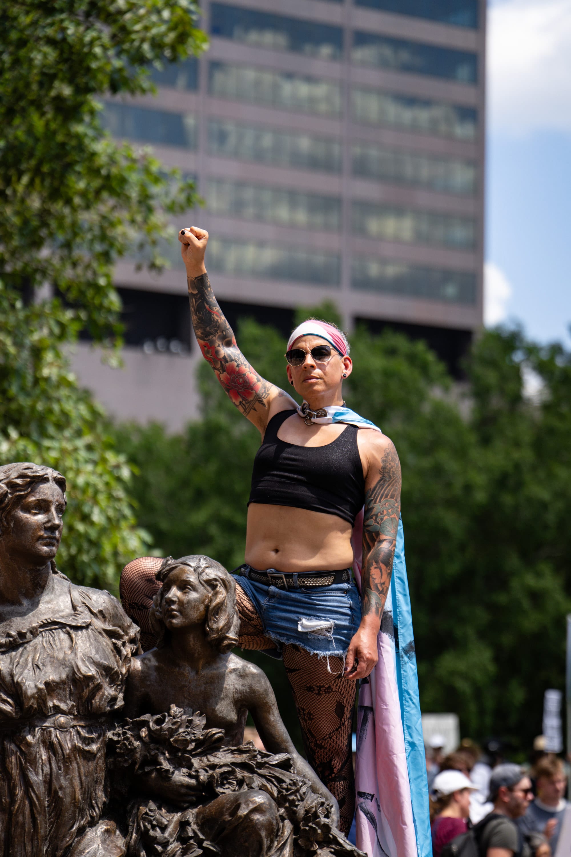 A woman raises a fist from beside a bronze statue group, a pride flag draped over her shoulders.