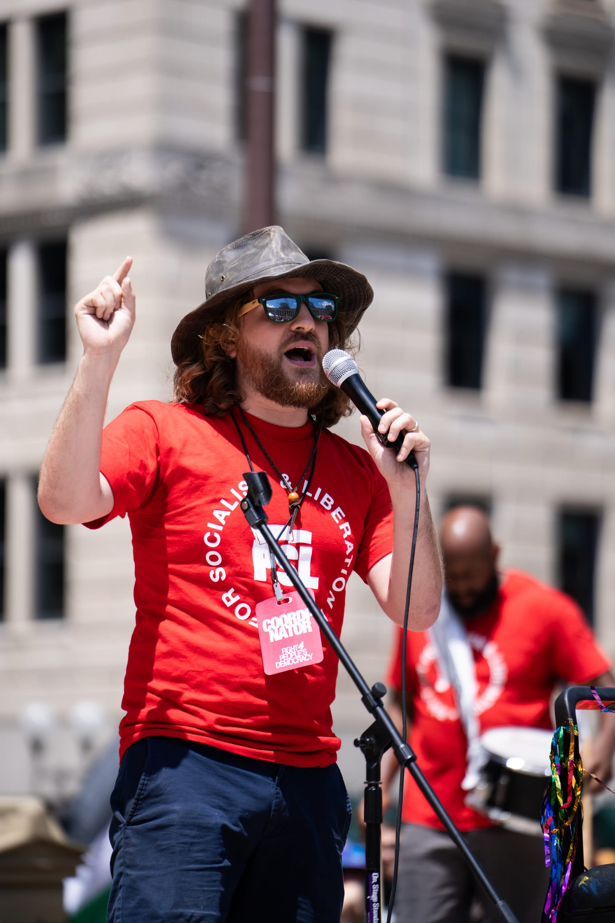 A bearded speaker in a bucket hat and red shirt points upward as he speaks into the microphone.