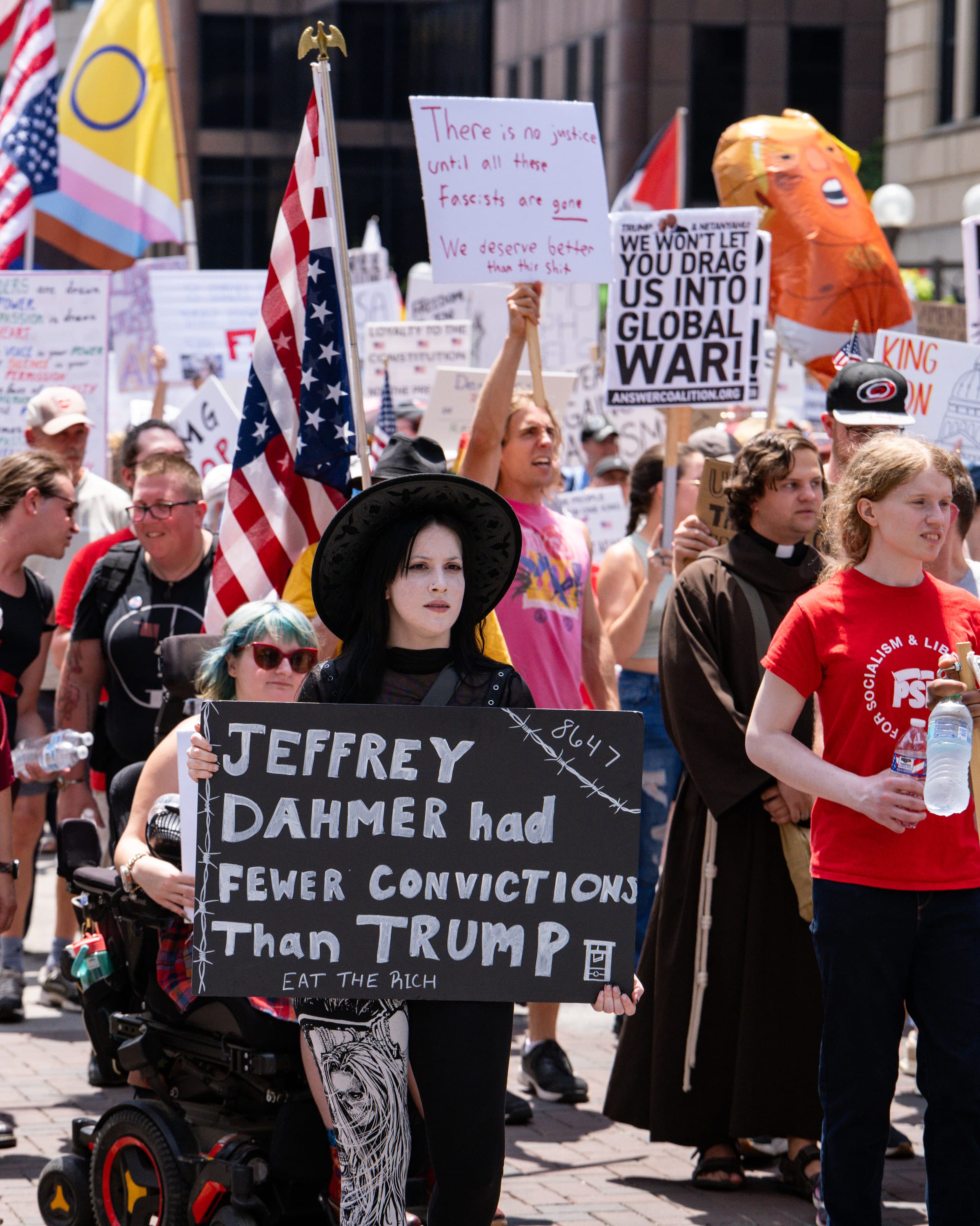 A marcher in a dark wig holds a chalkboard sign reading 'Jeffrey Dahmer had fewer convictions than Trump', flags and signs all around.