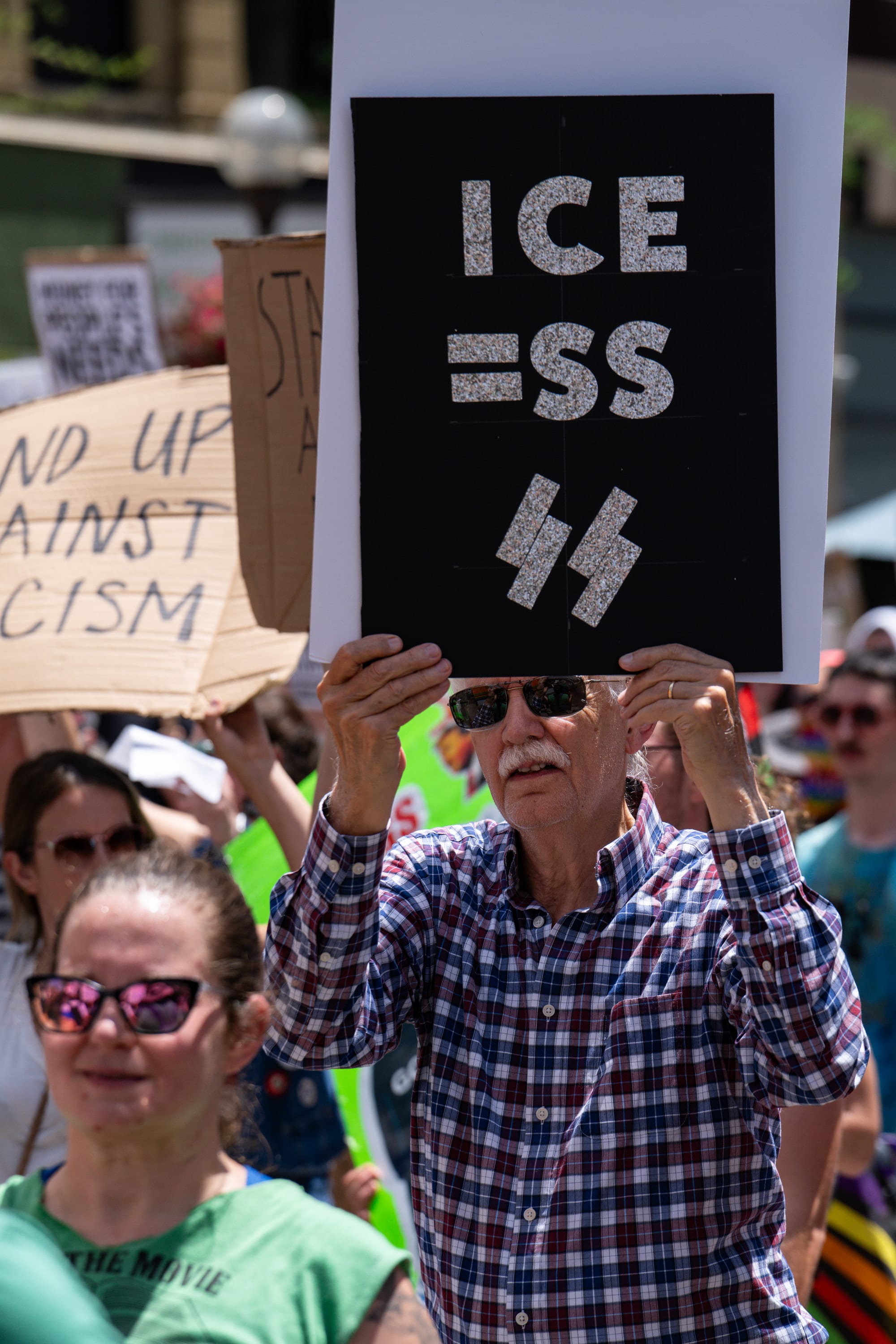 An older man holds up a black sign reading 'ICE = SS' with SS runes.
