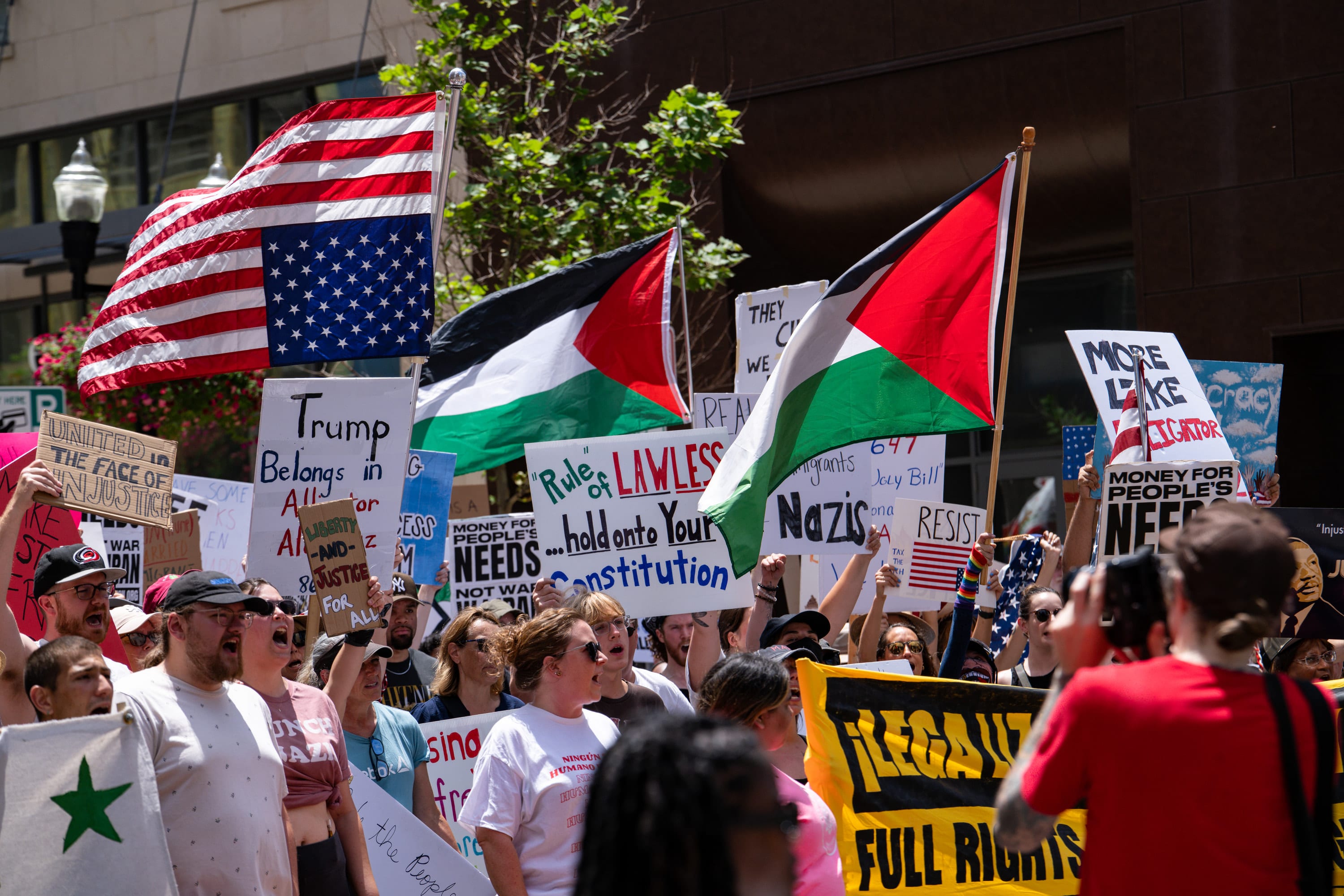 Palestinian flags and an upside-down American flag wave over a crowd holding 'Trump belongs in Alcatraz' and 'Hold on to your Constitution' signs.