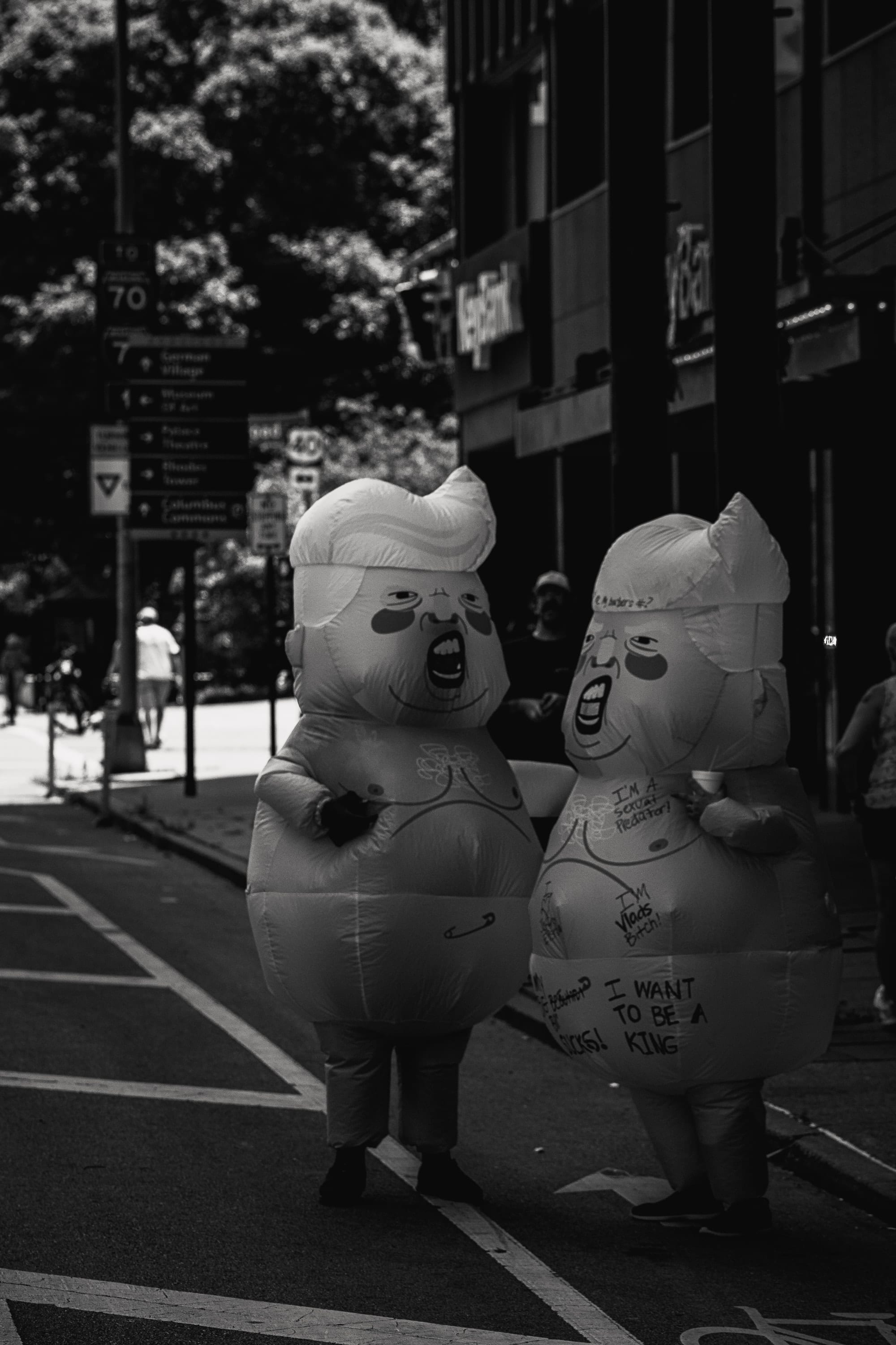 Black and white: two inflatable baby-Trump costumes walk down an empty street.