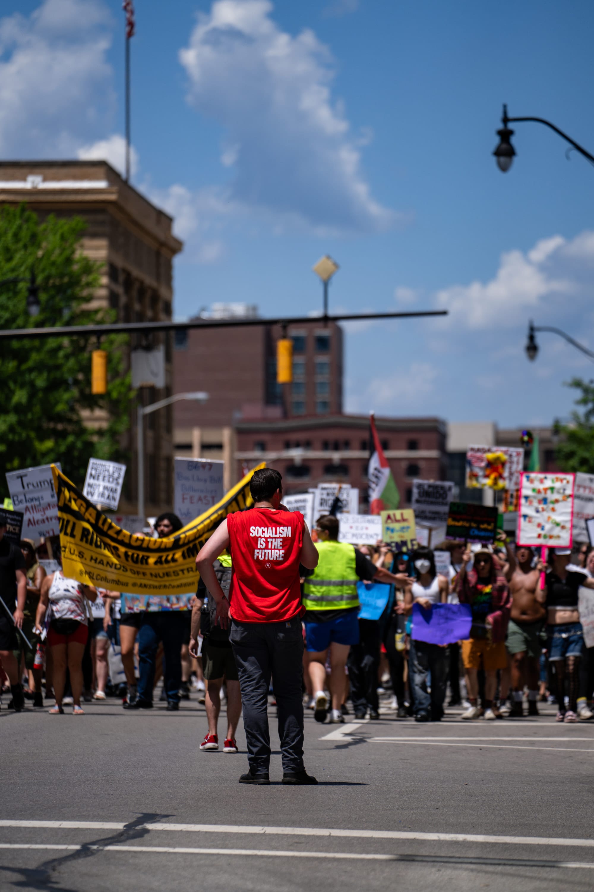 A marshal in a red shirt stands in the intersection as the march approaches under a blue sky.