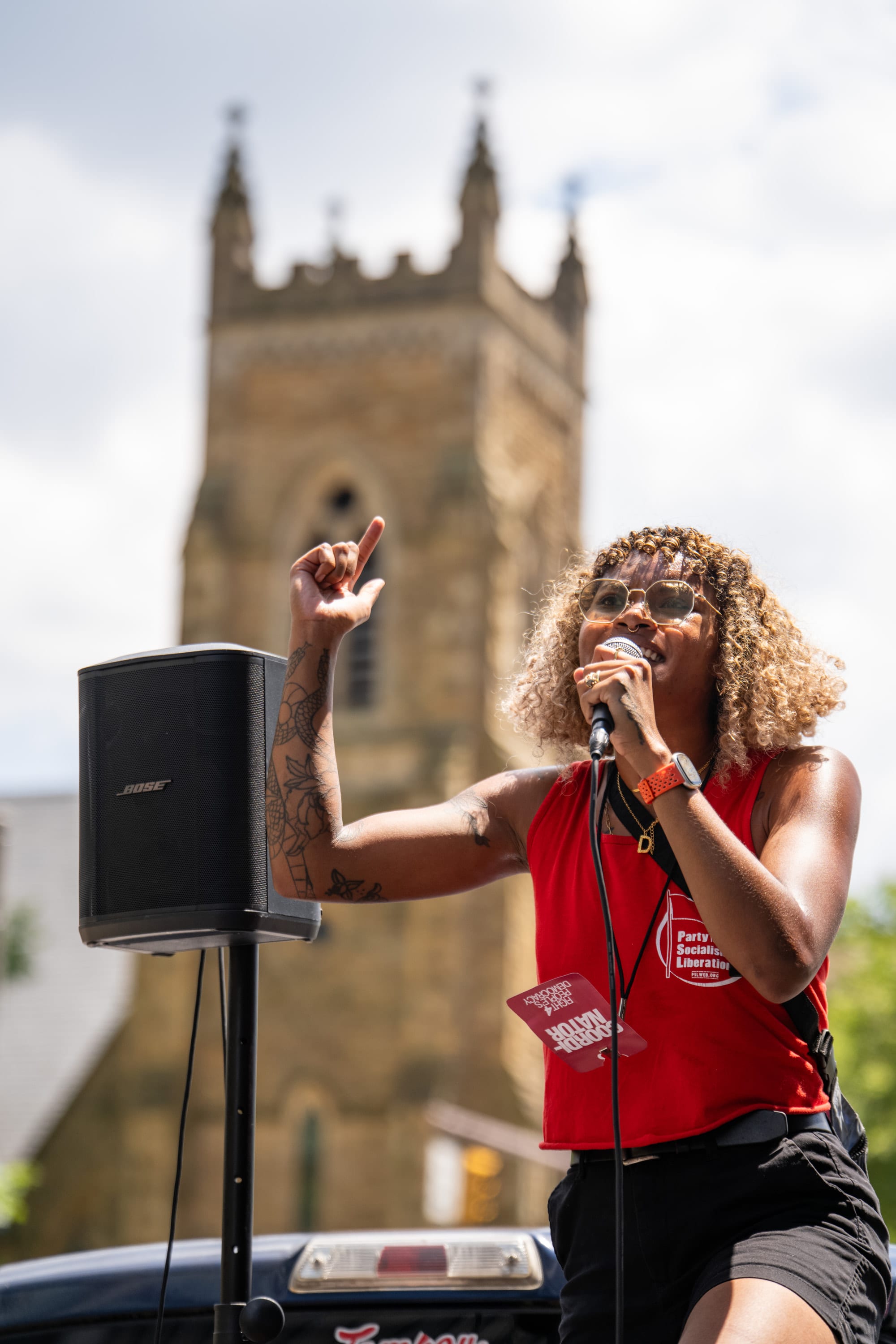 A speaker in a red top calls out into the microphone beside a speaker stack, a church tower rising behind her.