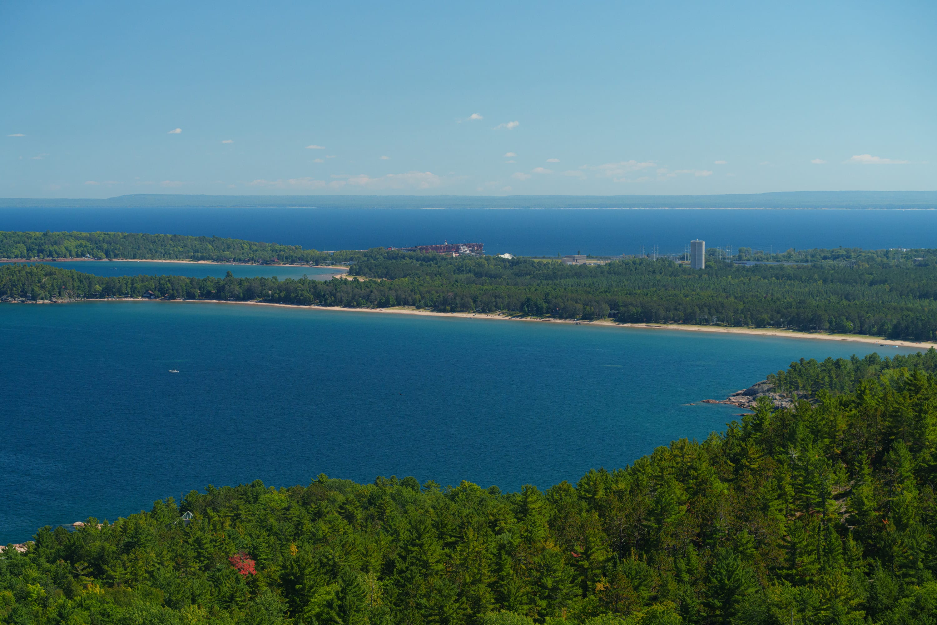 A curving sand beach and forested shore along a vast blue lake, a town with a tall building far off.