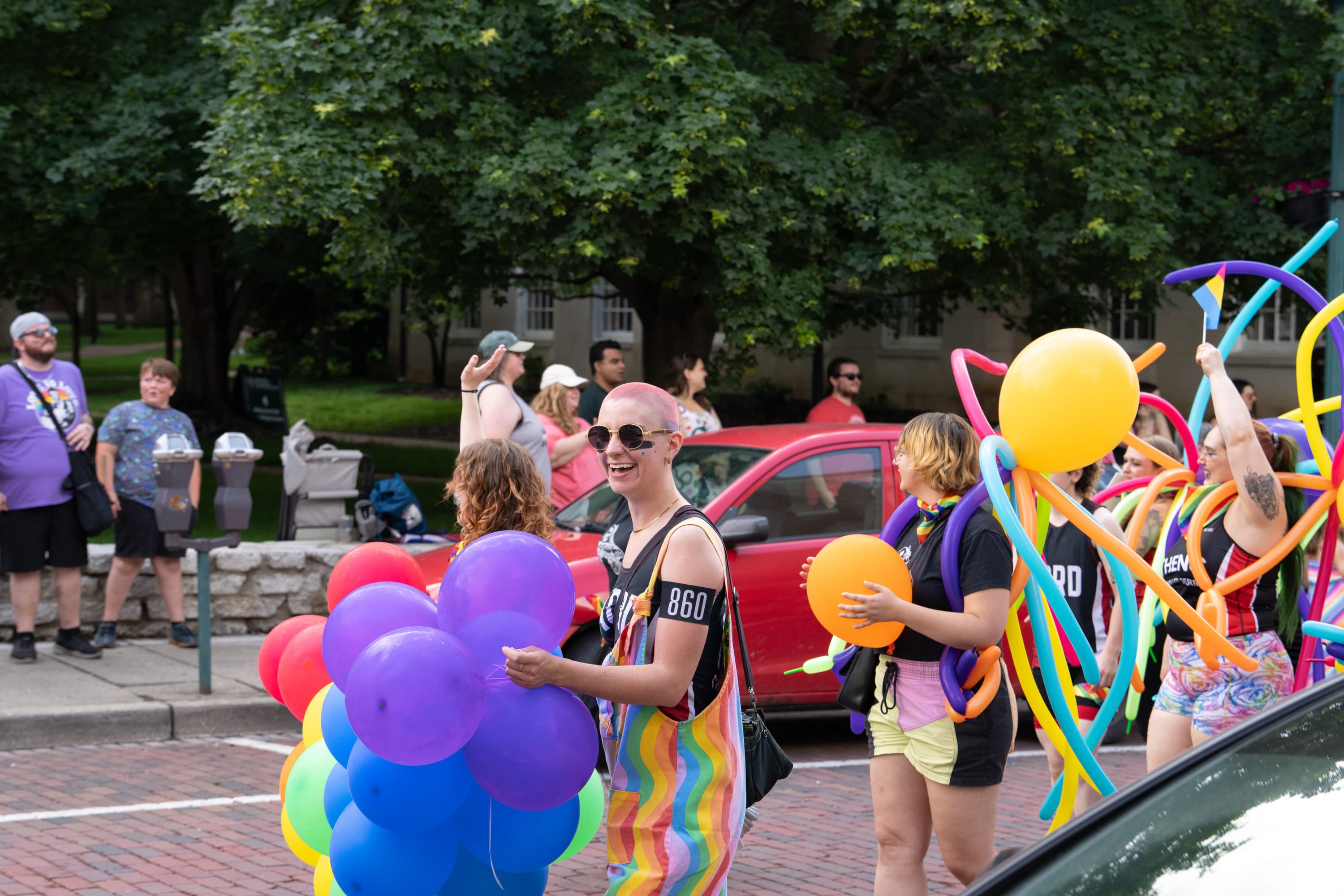 Parade marchers carry bunches of rainbow balloons down a brick street, one in rainbow overalls.