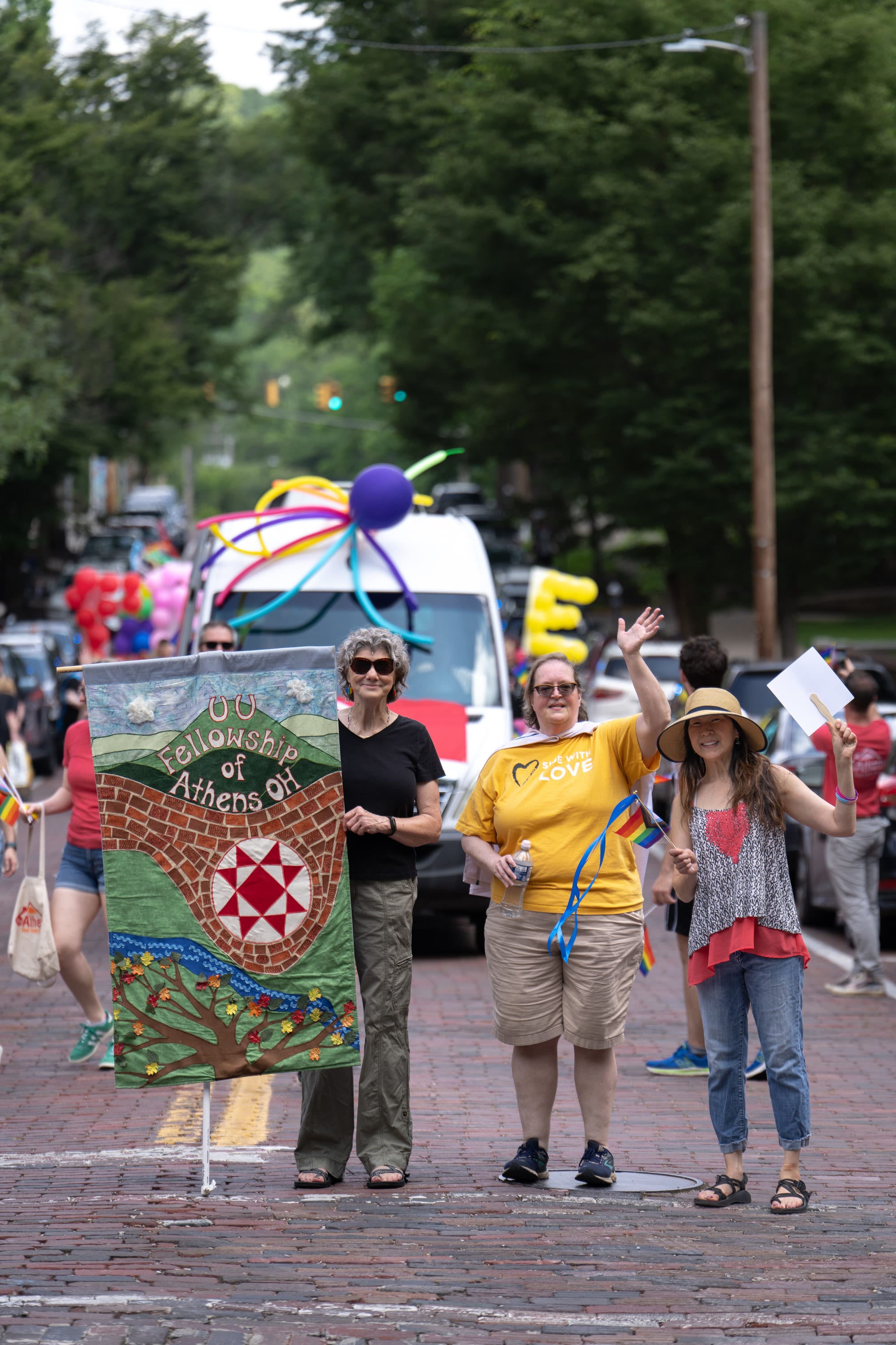 Marchers smile and wave behind a quilted banner on a brick street.