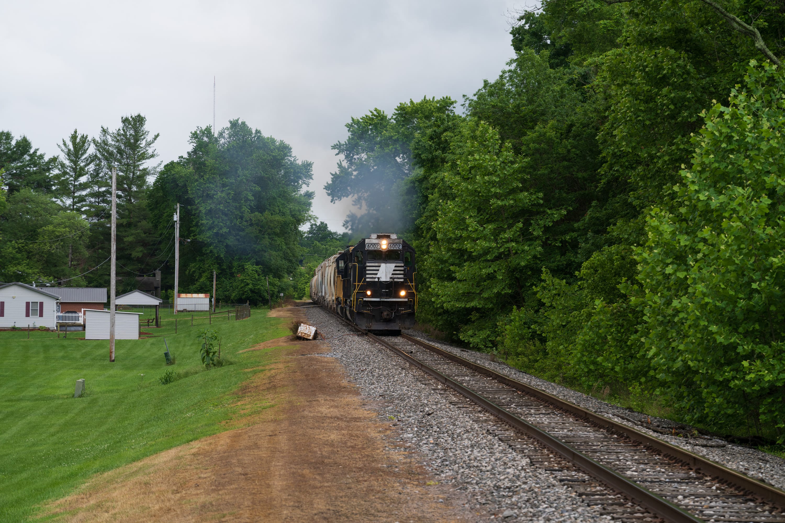 A freight train approaches past a lawn and white houses, exhaust rising above the trees.