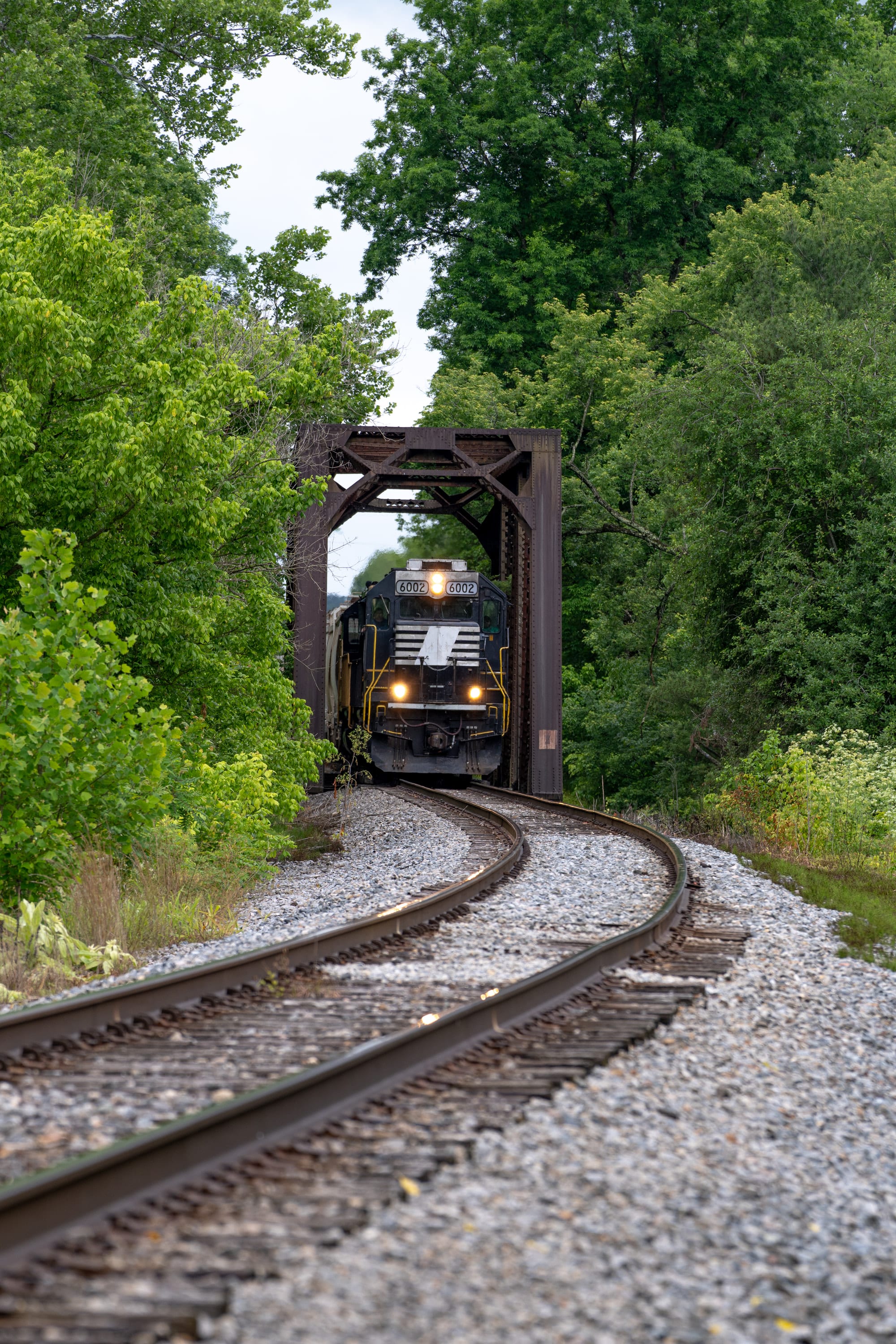 A locomotive crosses a steel truss bridge, framed by green trees and curving rails.