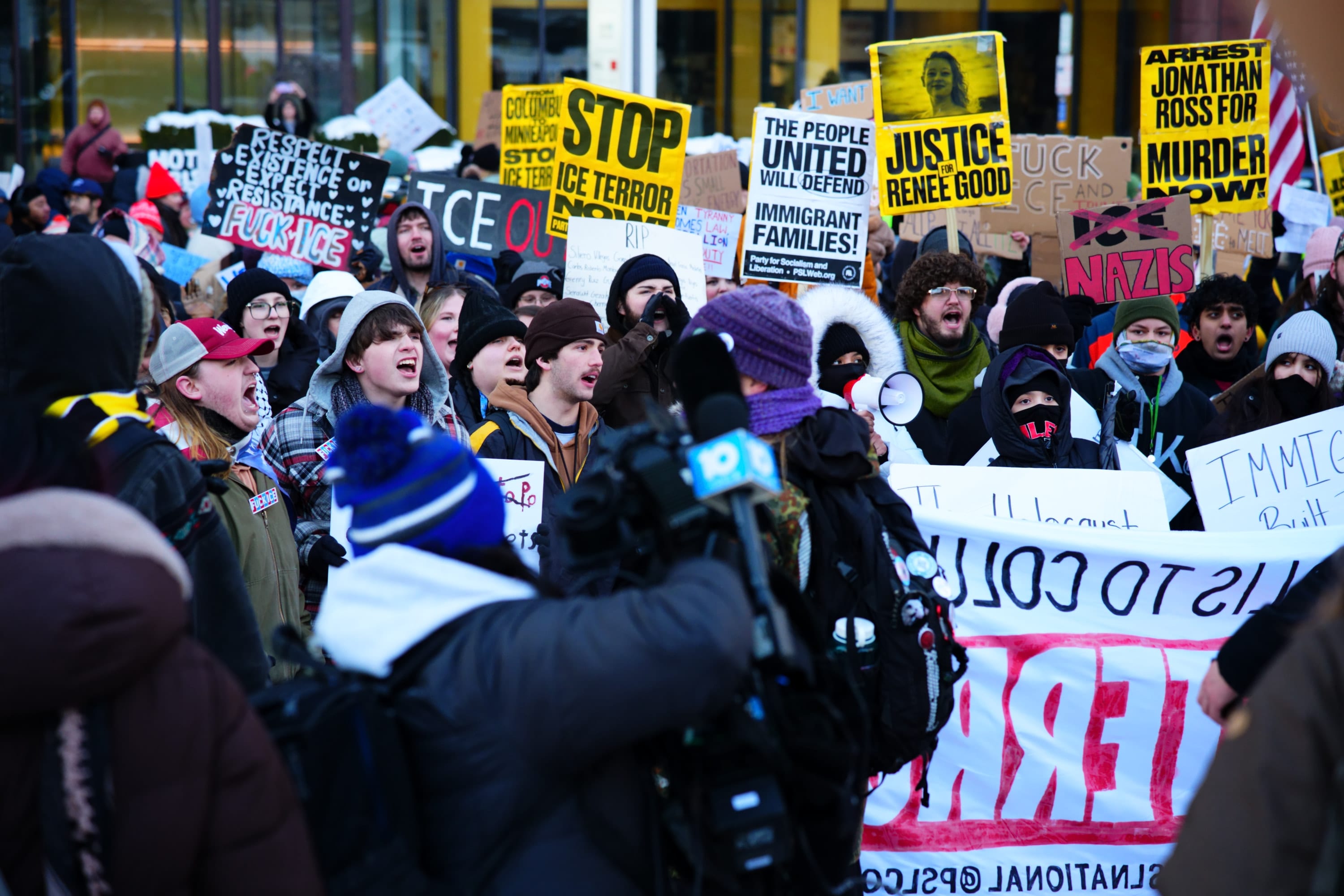 A crowd packed shoulder to shoulder behind 'Stop ICE terror' and 'The people united will defend immigrant families' signs as a TV camera films.