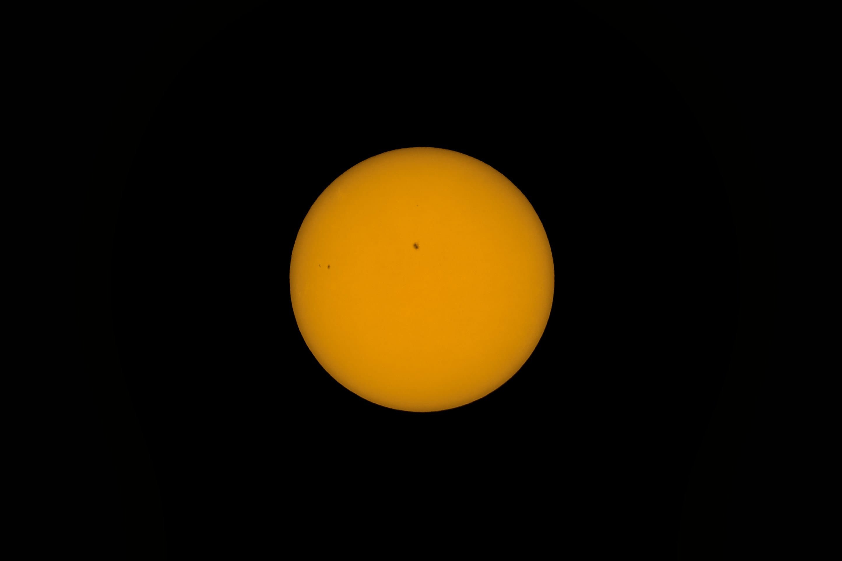 The sun through a solar filter, an orange disc on black with one small dark sunspot.