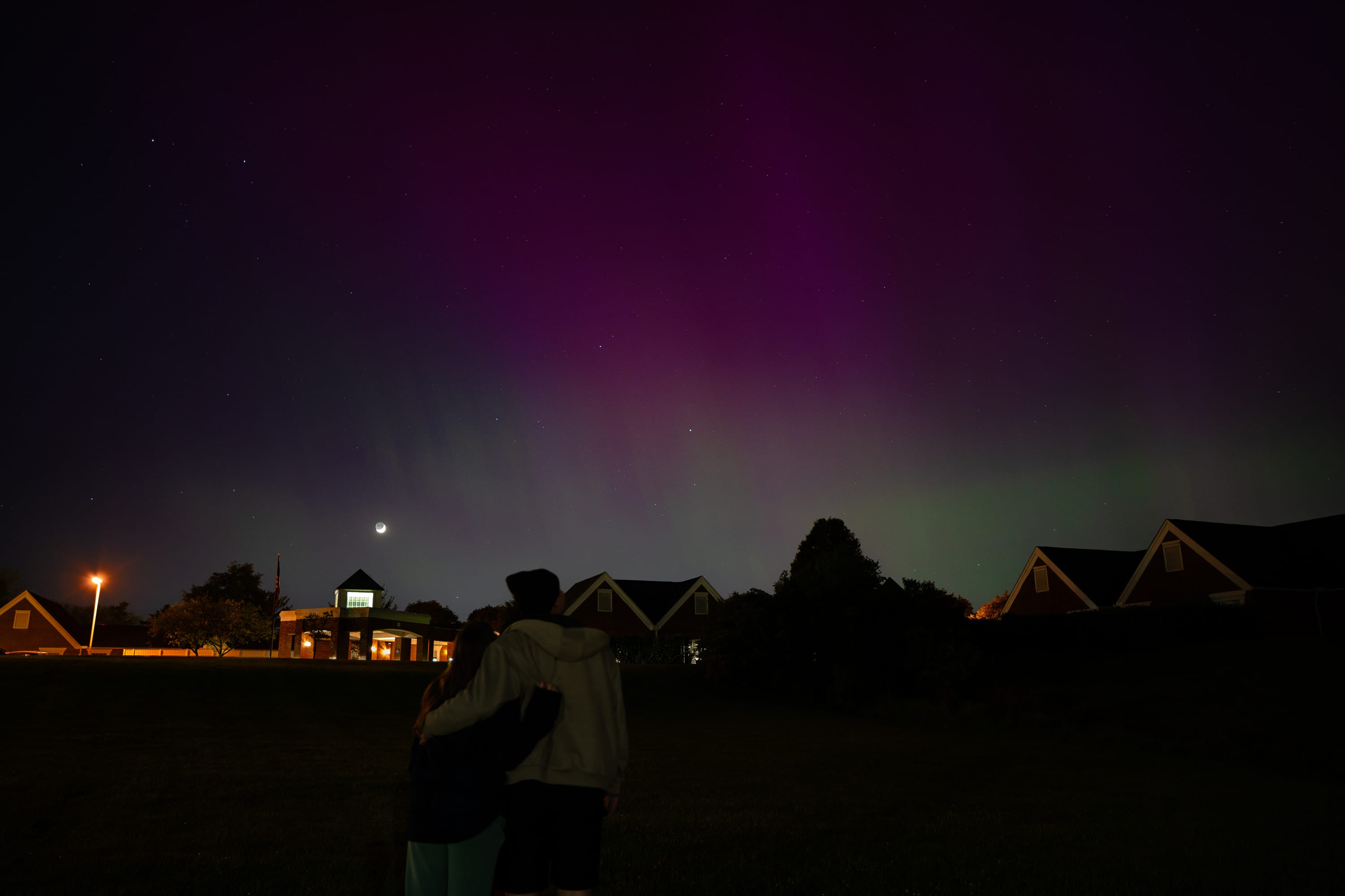 Magenta and green aurora over a row of houses at night, two figures silhouetted in the foreground.