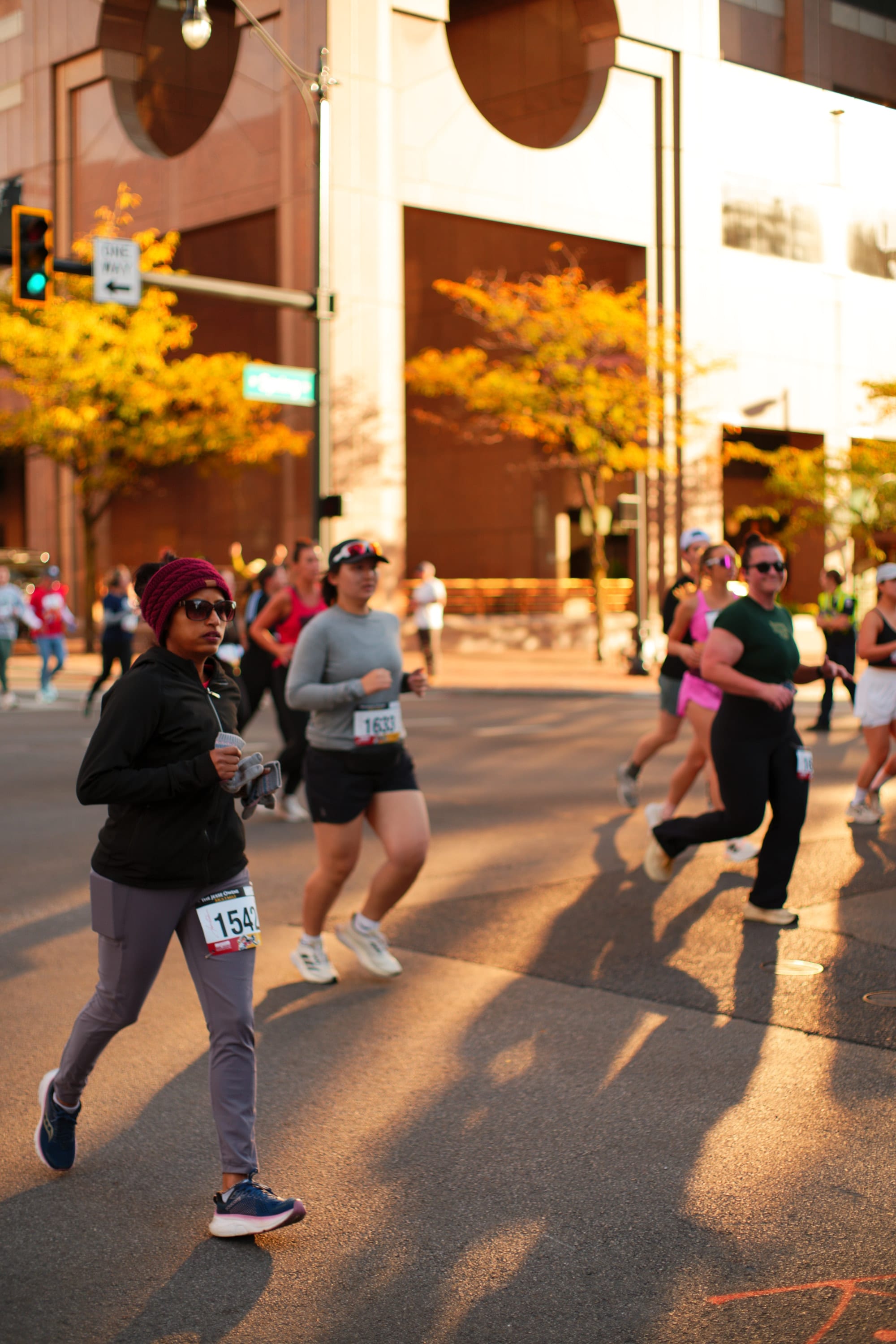 Runners in race bibs stream down a downtown street at sunrise, long shadows stretching ahead of them.